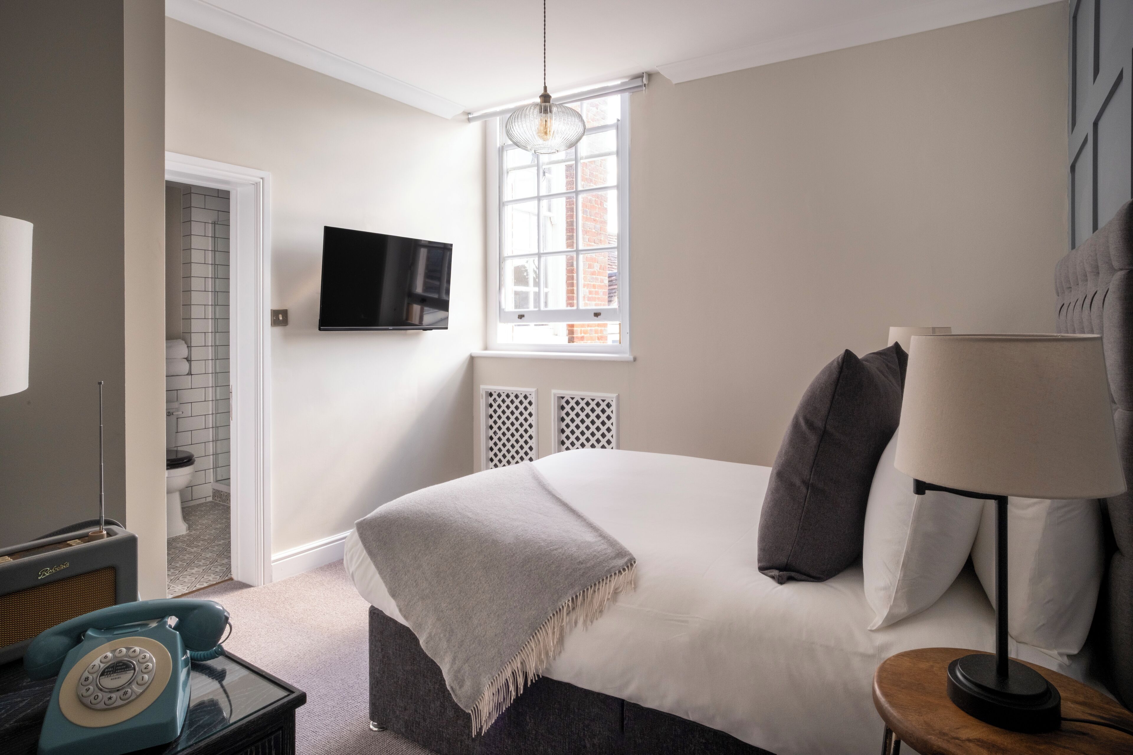 Un dormitorio moderno con una cama grande, un televisor montado en la pared, una ventana con vistas a un edificio de ladrillo y un teléfono antiguo sobre una mesita de noche.