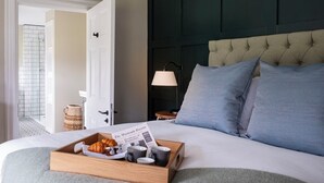 Et hotelværelse med en pænt redt seng, et bakke med morgenmad, en avis og udsigt til et badeværelse med en flisebeklædt brusekabine.