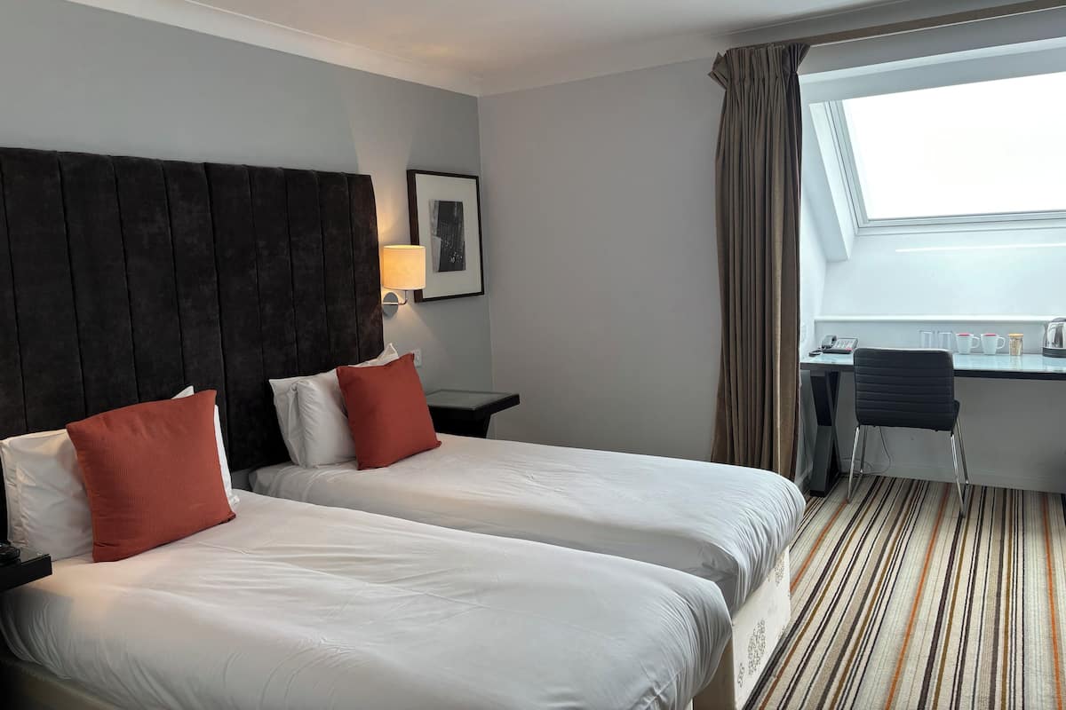 Habitació d'hotel amb dos llits, escriptori, cadira, làmpada i finestra amb cortines.