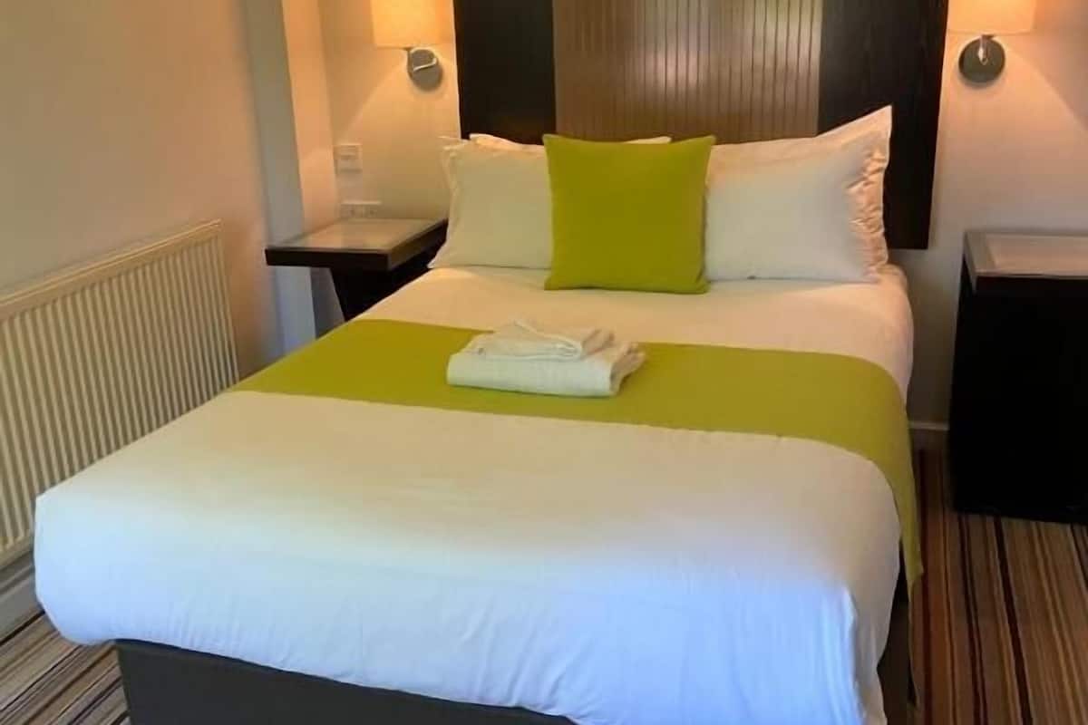 Habitació d'hotel amb un llit gran, un capçal amb ratlles verticals, dues taules de nit i una tovallola doblegada sobre el llit.