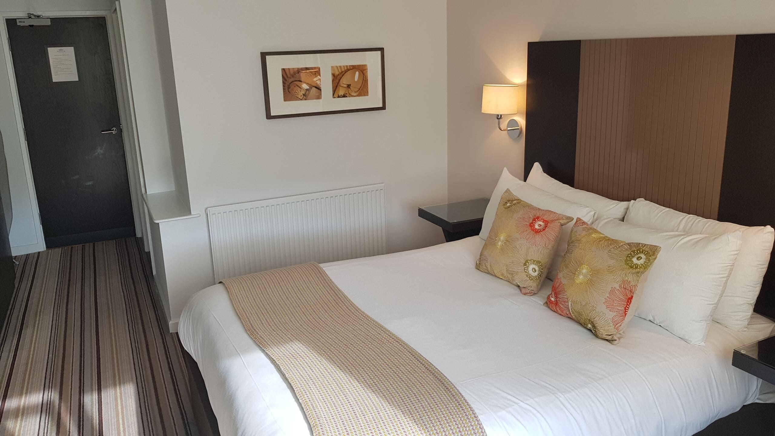 A hotel room with a large bed, a bedside table, a wall-mounted lamp, and a framed picture on the wall.