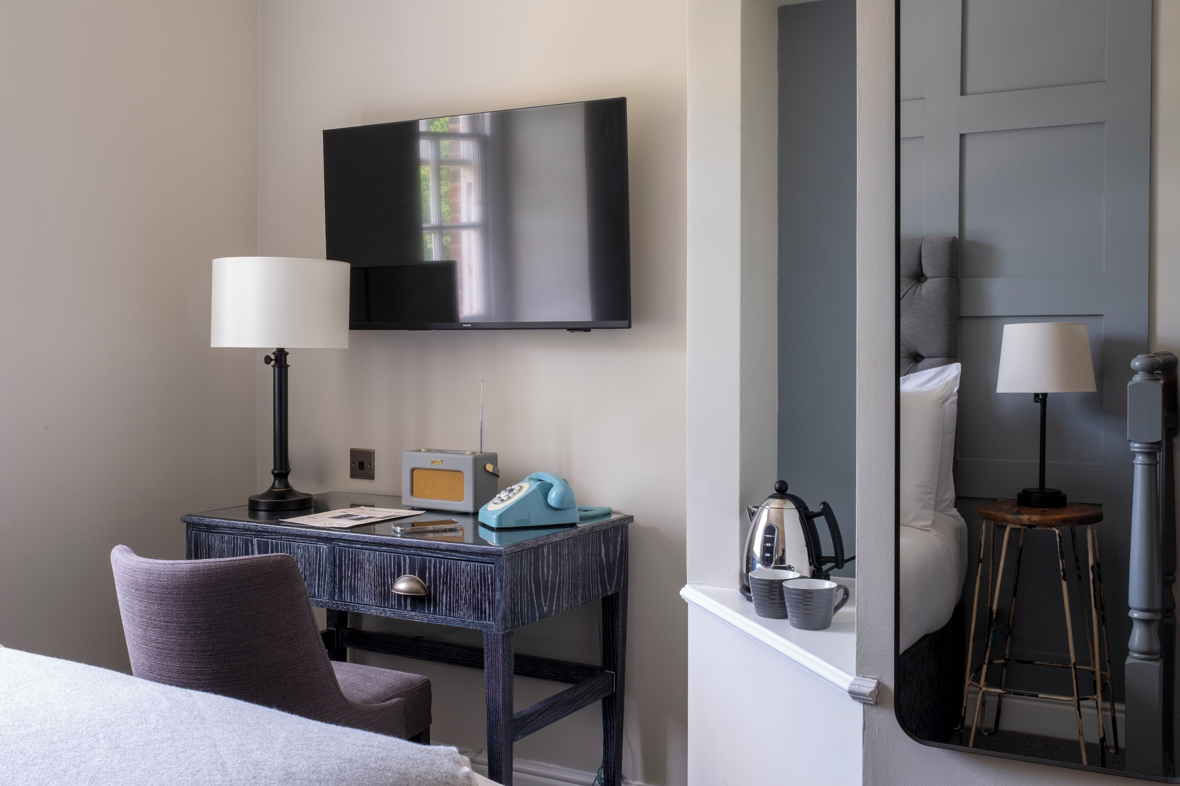 A hotel room with a bed, a desk with a lamp, a television, a chair, a small table with a kettle, and a mirror.