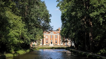 A river with ducks, surrounded by trees, leading to a red brick building with multiple windows and a central entrance.