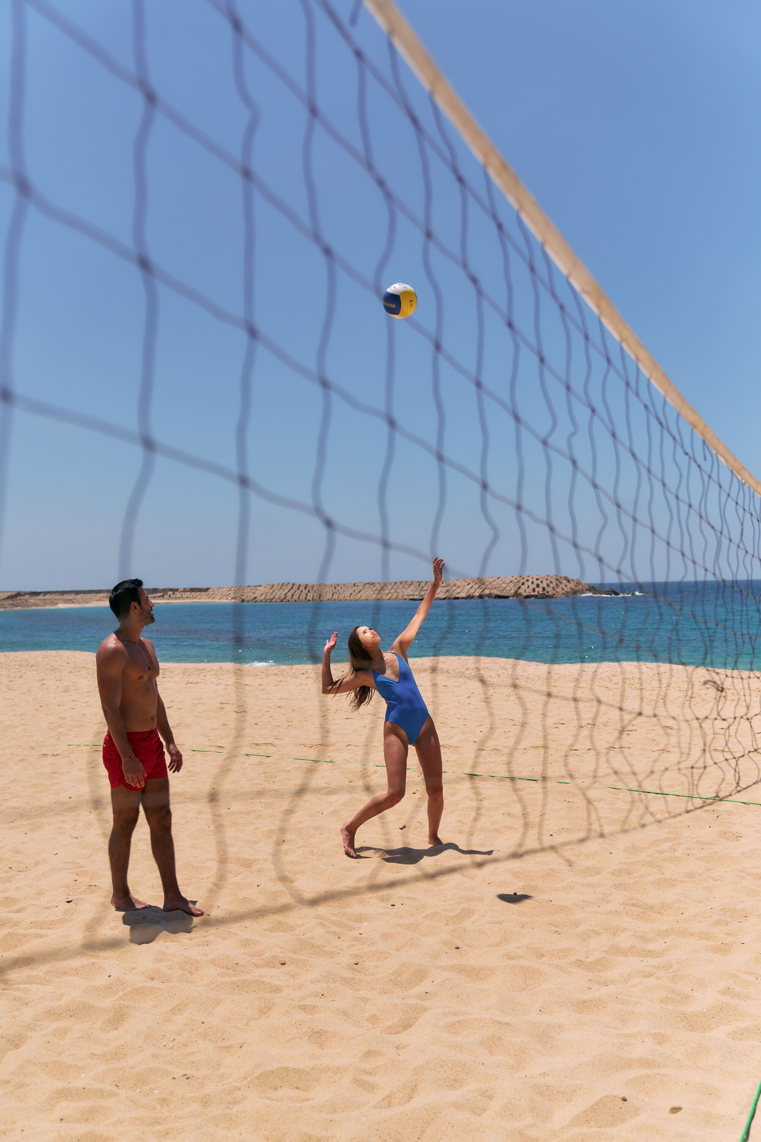 A beach volleyball game with a net and players on a sandy beach with a clear sky and ocean in the background.