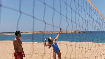 A beach volleyball game with a net and players on a sandy beach with a clear sky and ocean in the background.