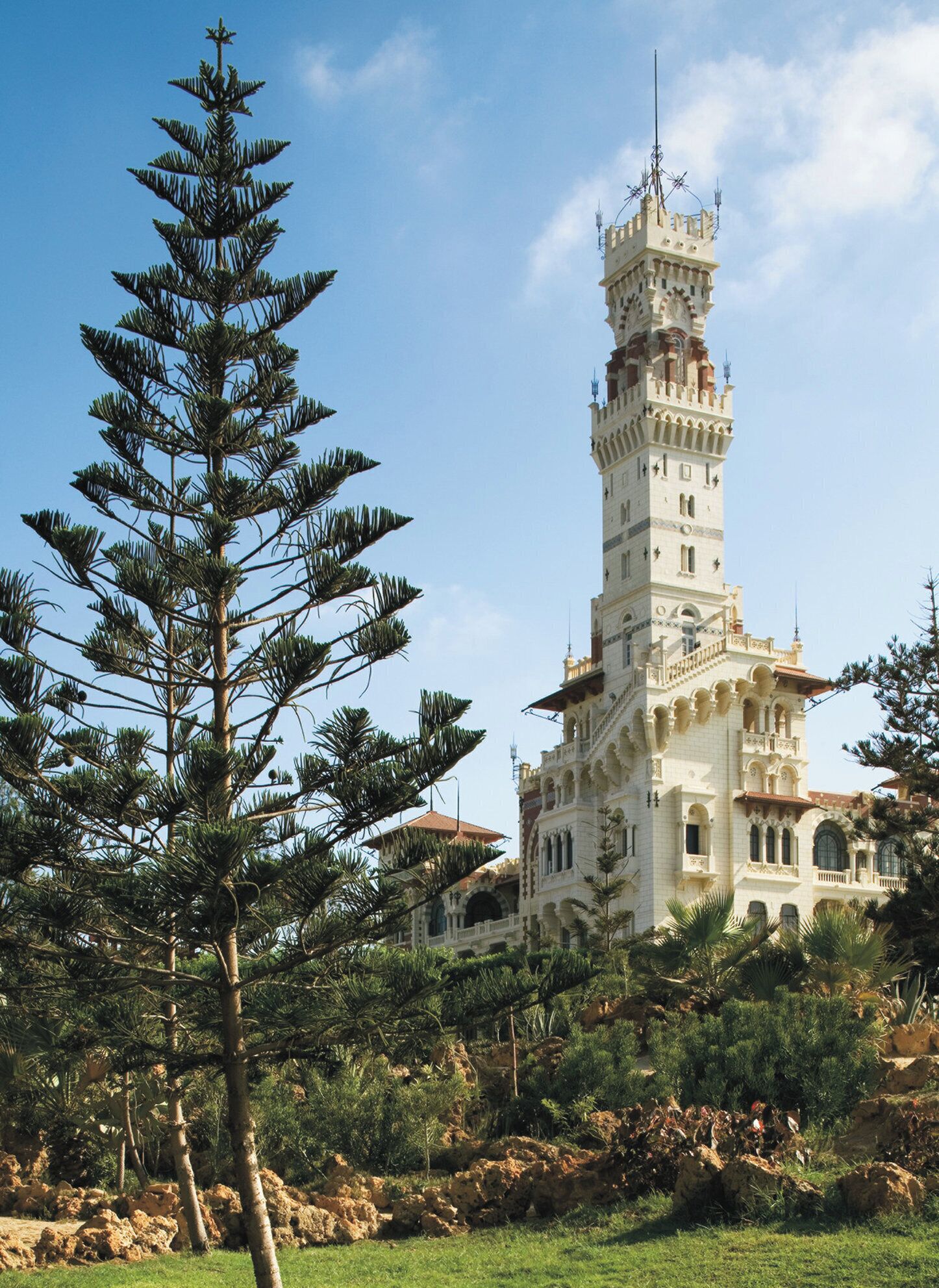 A tall, ornate tower with intricate architectural details, surrounded by lush greenery and a clear sky.