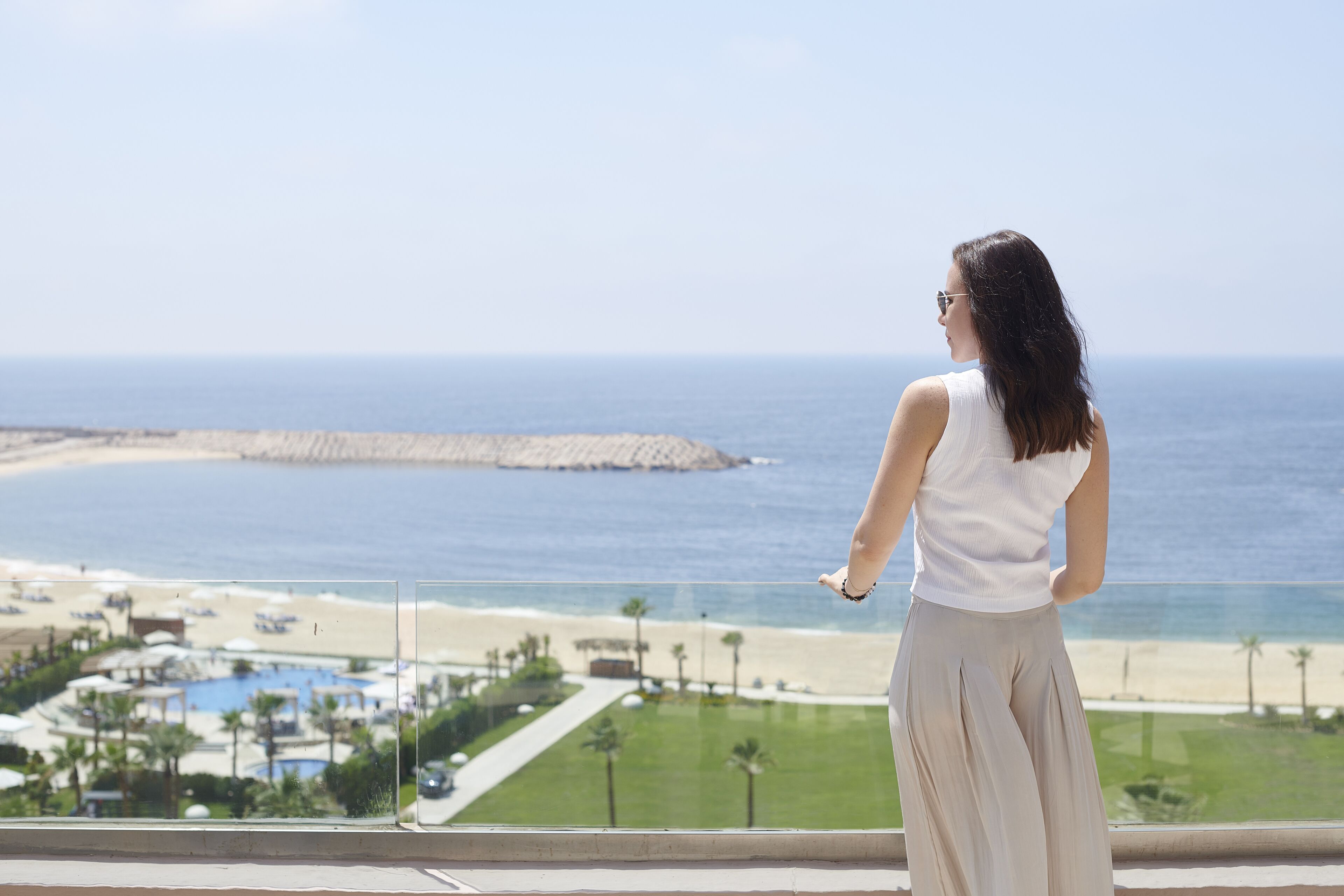 A woman standing by a balcony overlooking a beach with a pool and palm trees.