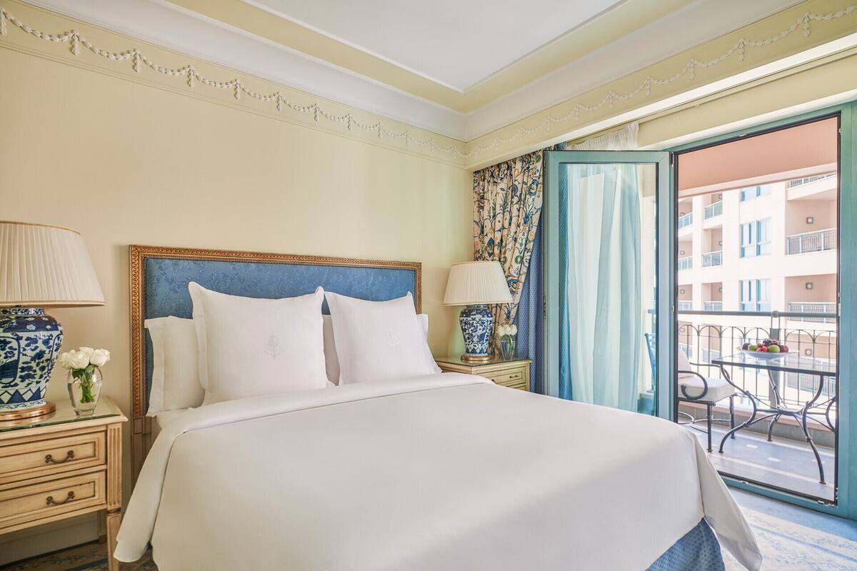 A hotel room with a large bed, two bedside tables with lamps, a view of a balcony with outdoor furniture, and a patterned wallpaper on the wall.