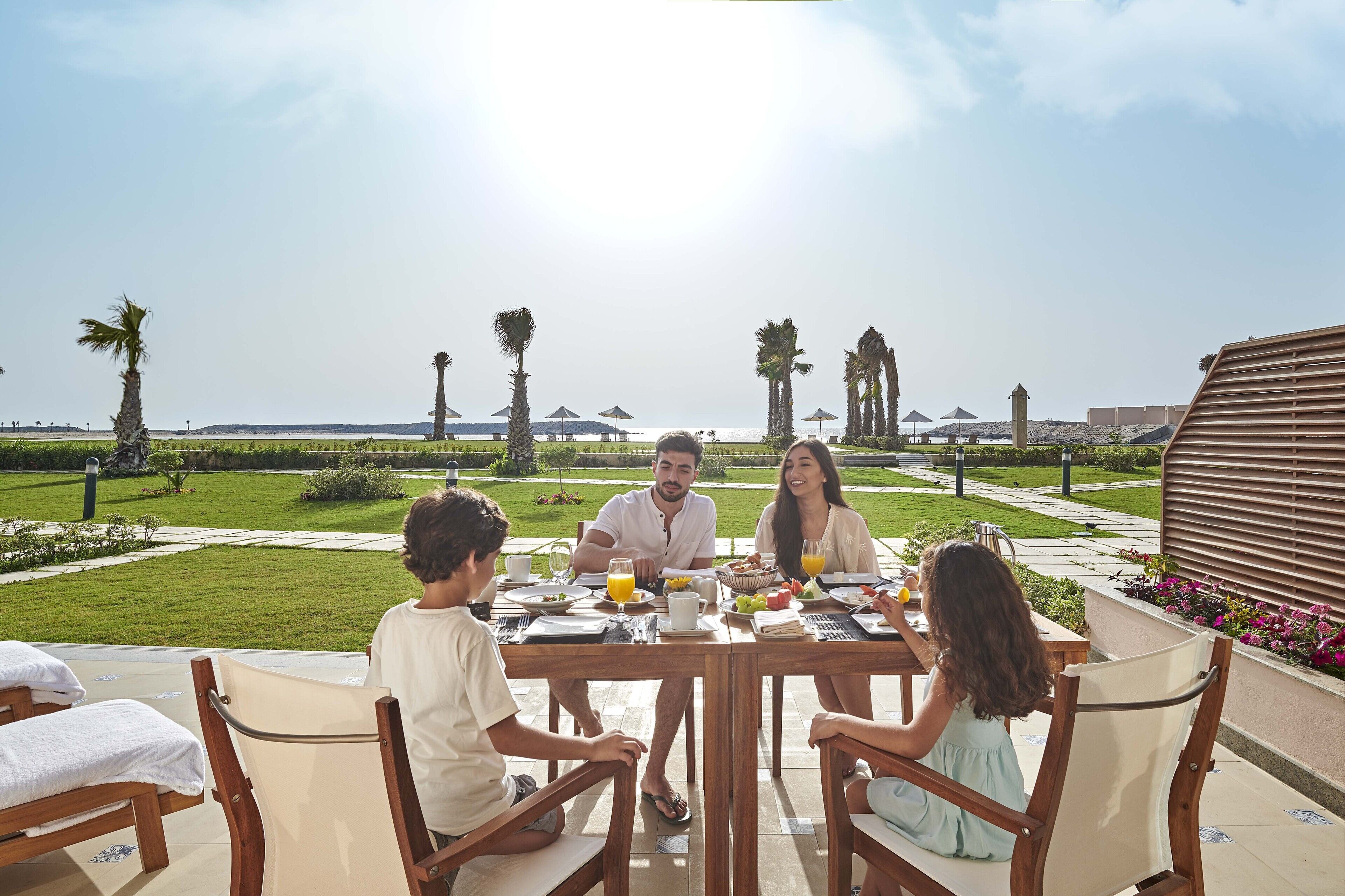 Una familia cenando en una mesa al aire libre con vista al mar y palmeras.