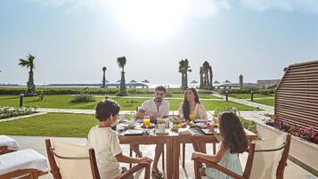 Una familia cenando en una mesa al aire libre con vista al mar y palmeras.