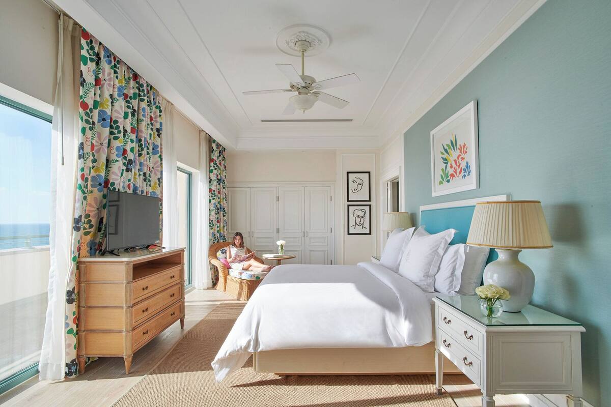 A hotel room with a bed, a dresser, a nightstand, a ceiling fan, and a large window with curtains.