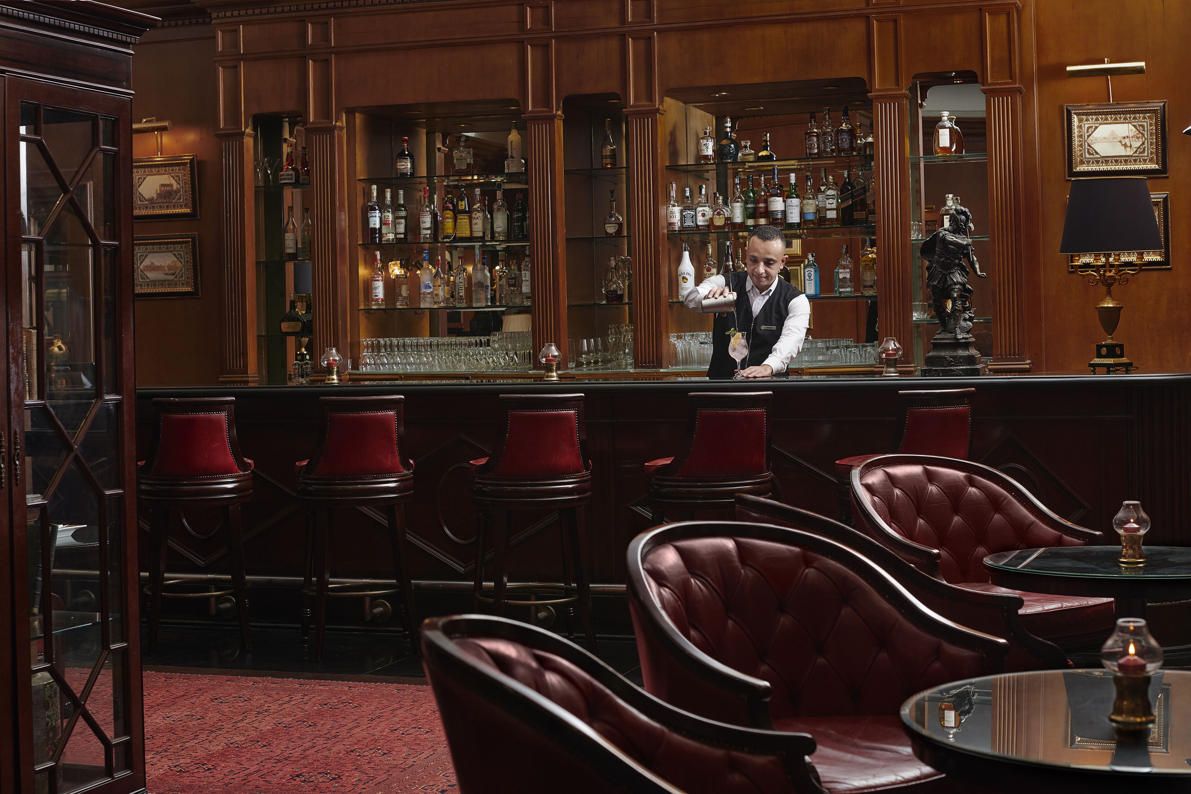 A bar area with leather chairs, a bartender, and a well-stocked liquor shelf.