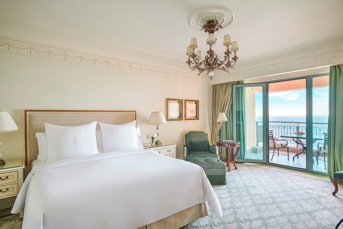 A hotel room with a large bed, a chandelier, a balcony with a table and chairs, and a view of the sea.