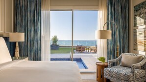 Habitación de hotel con una cama grande, una silla, una mesita y vista al océano a través de una puerta corredera.