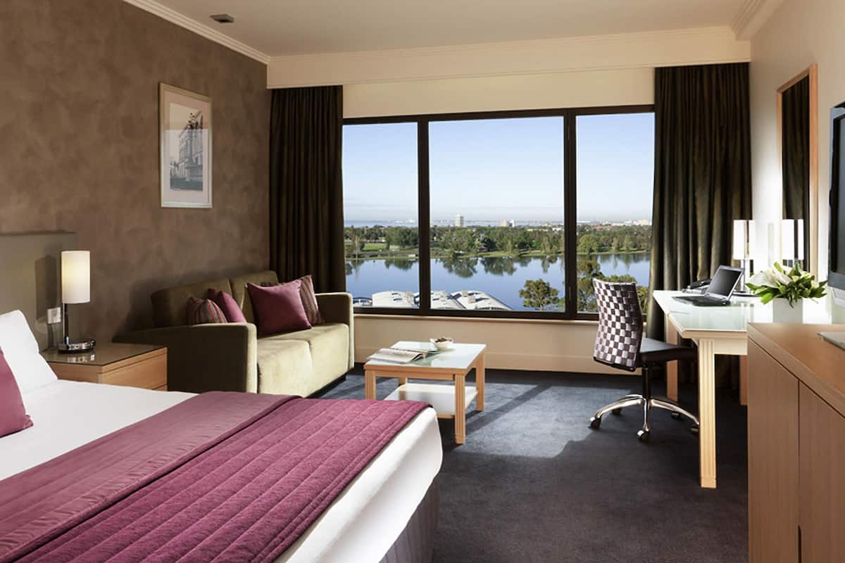 A hotel room with a large bed, a desk, a chair, a sofa, and a view of a lake.