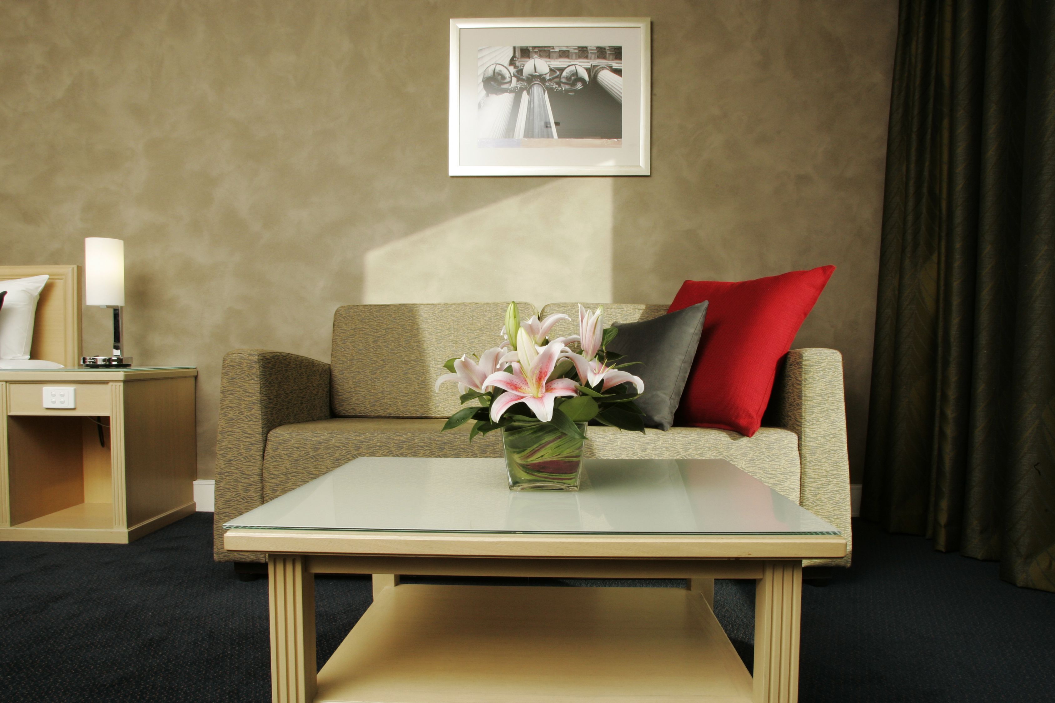 Een hotelkamer met een beige bank, een glazen bijzettafeltje, een vaas met bloemen, een lijst met een afbeelding aan de muur en een lamp op een nachtkastje.