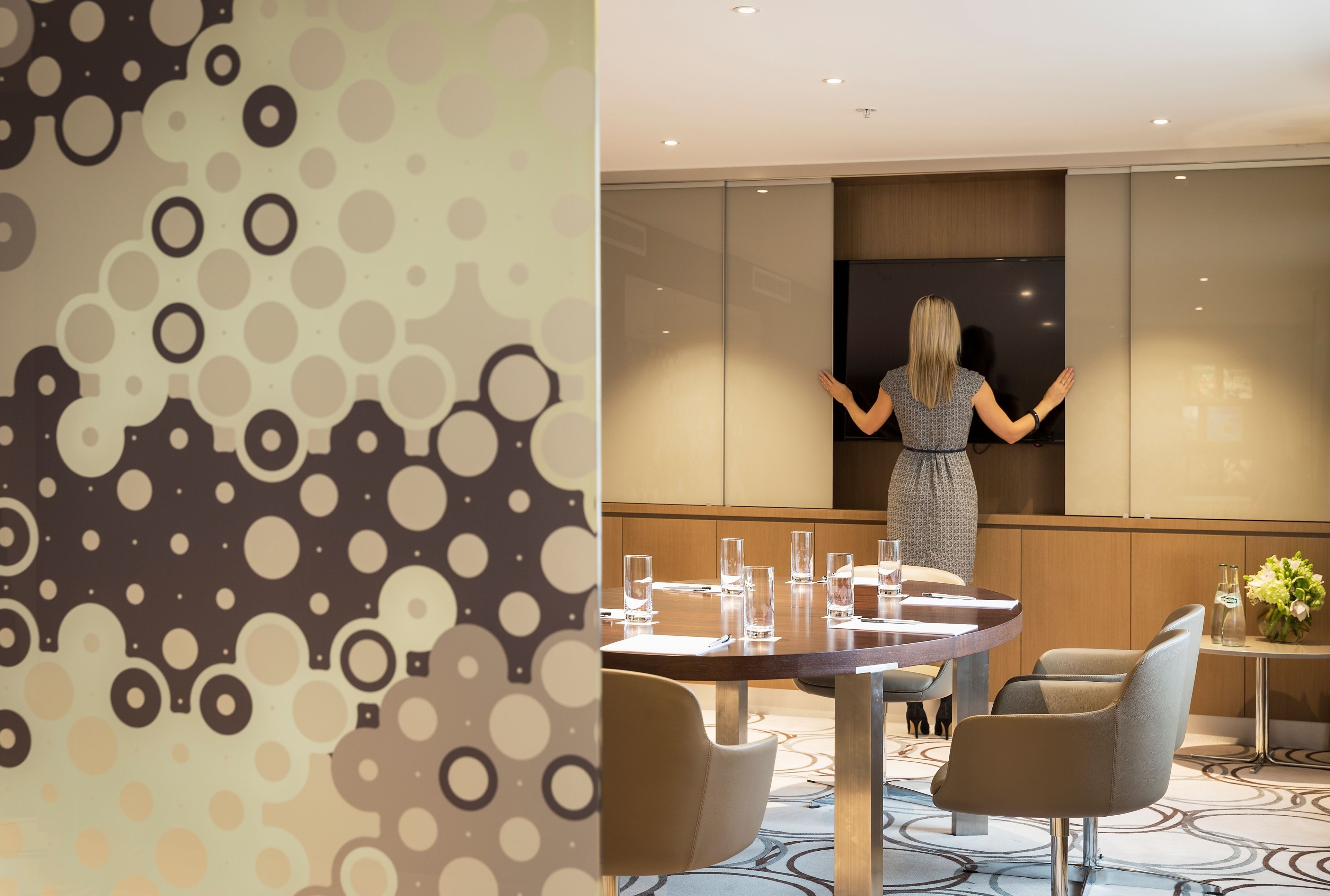 A conference room with a large screen, a round table with chairs, and a patterned wall.