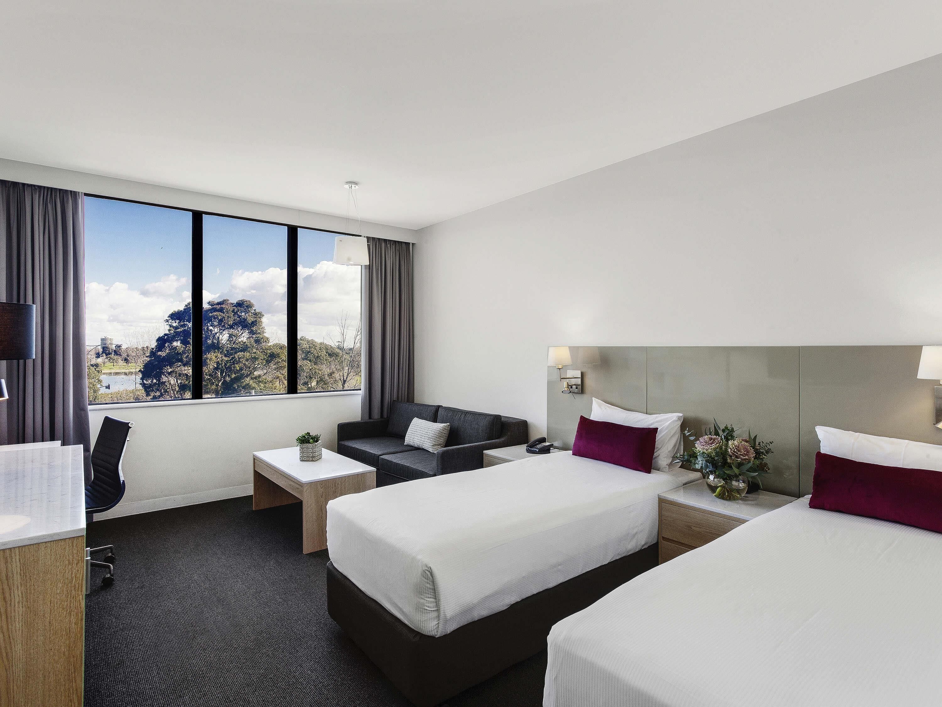 A hotel room with two beds, a desk, a chair, and a large window offering a view of trees and a body of water.