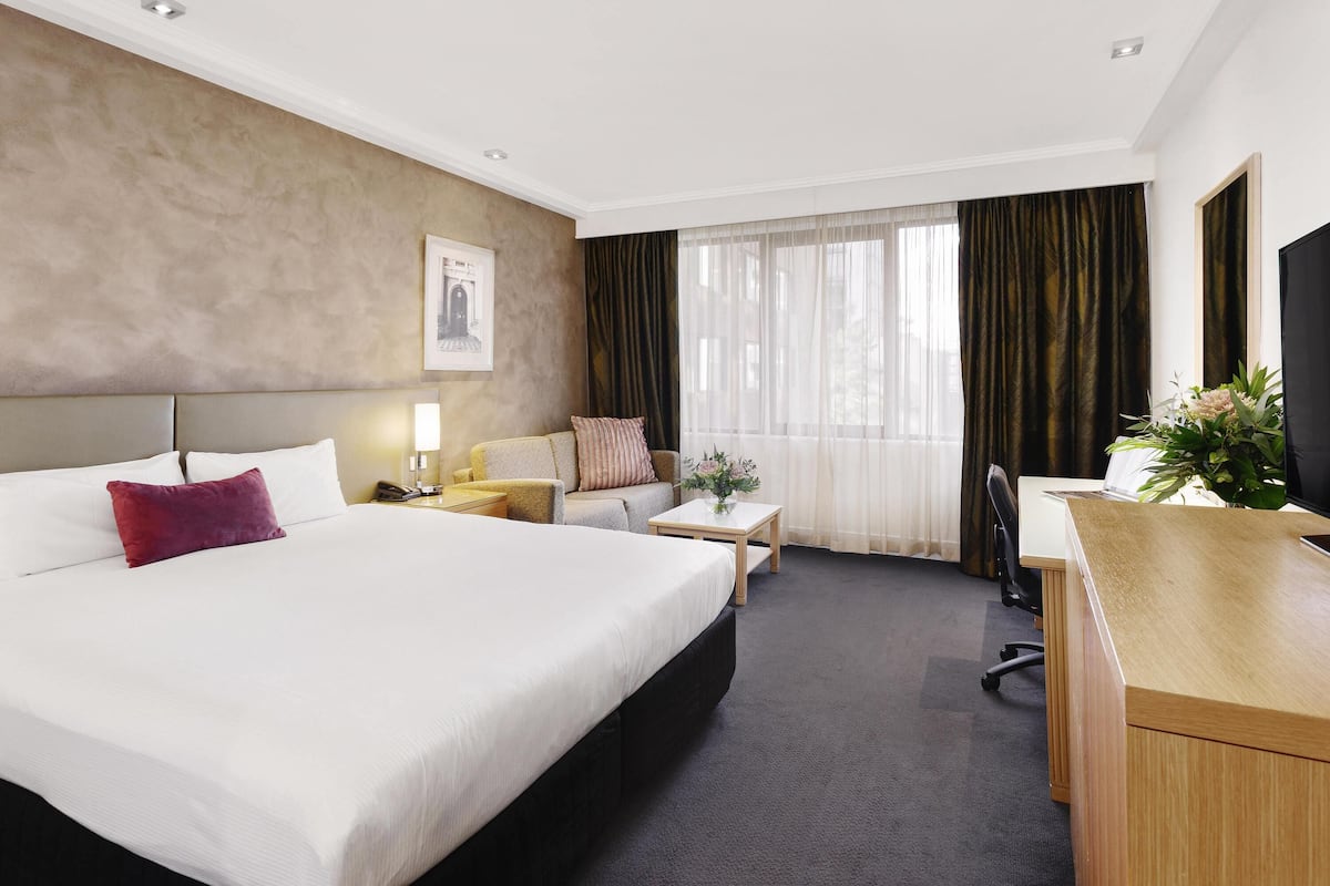 A hotel room with a large bed, a desk, a chair, a small table, a lamp, a TV, and a window with curtains.