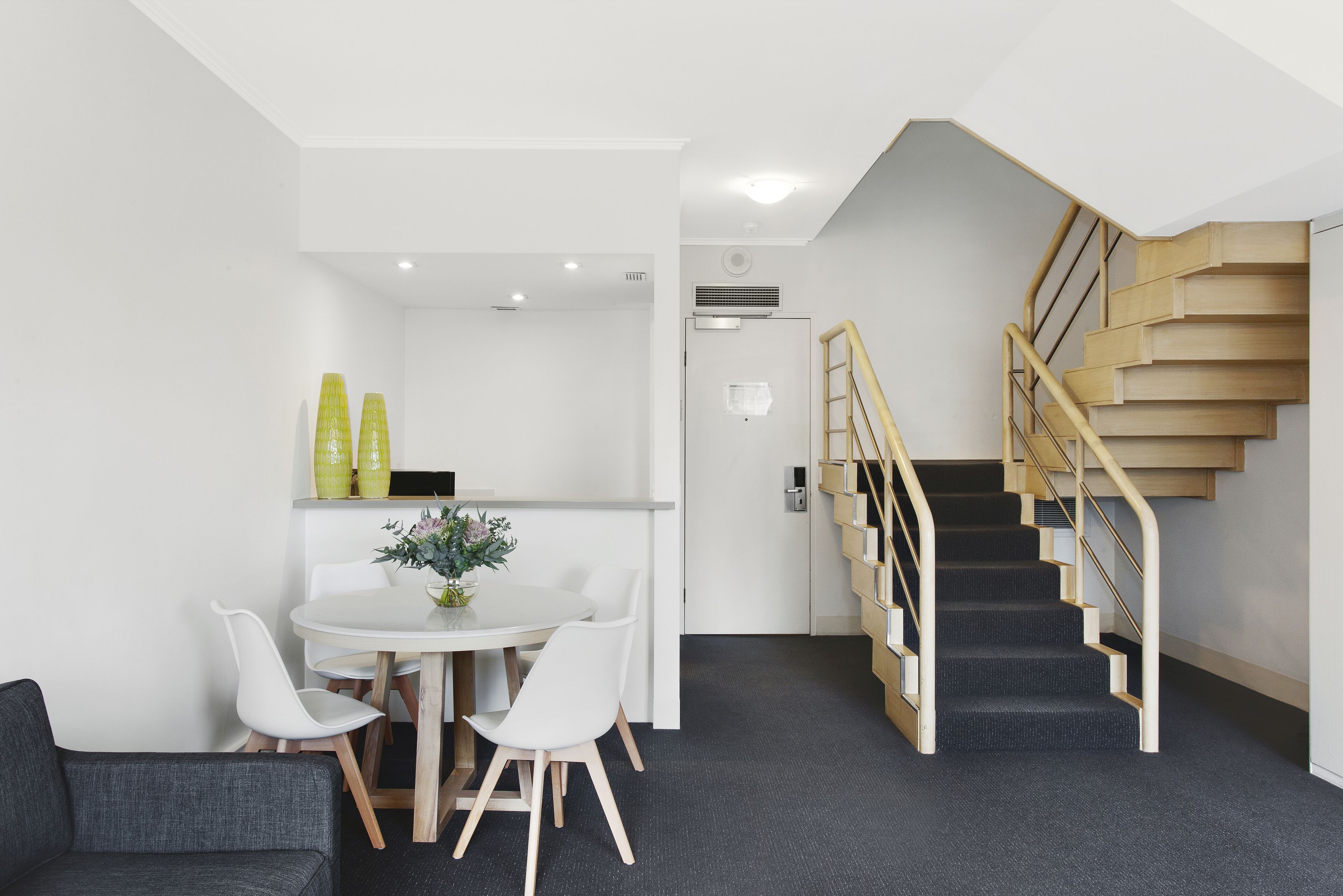 A modern interior with a staircase, a dining area with a table and chairs, and a grey carpeted floor.