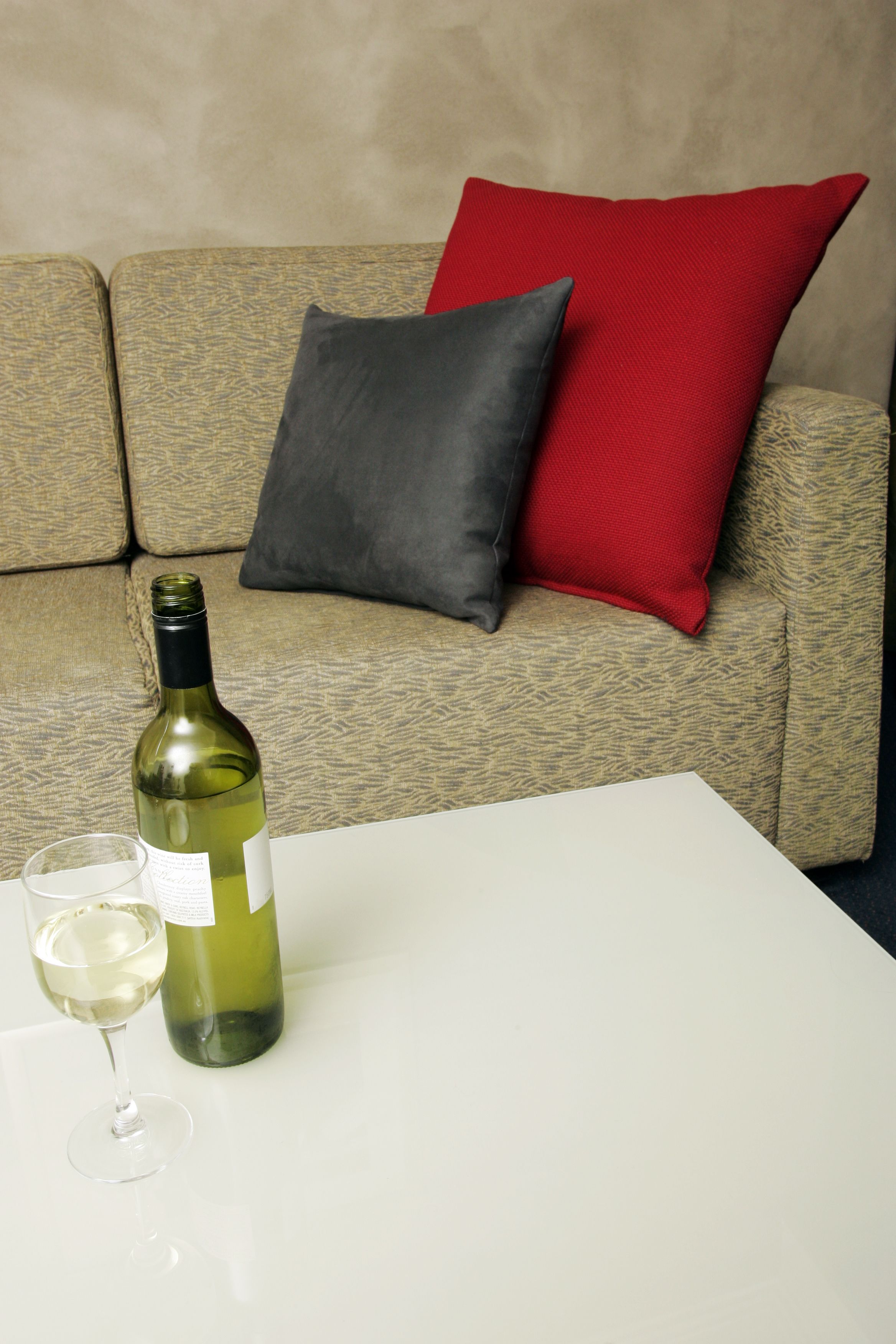 Une bouteille de vin blanc et un verre posés sur une table blanche devant un canapé beige agrémenté d’un coussin rouge et gris.