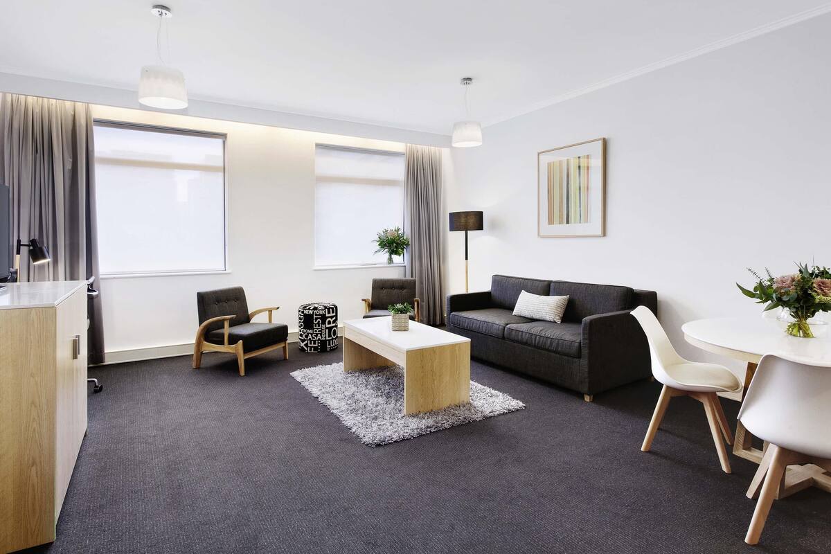 A modern living room with a sofa, coffee table, and chairs, a desk with a lamp, and large windows with curtains.