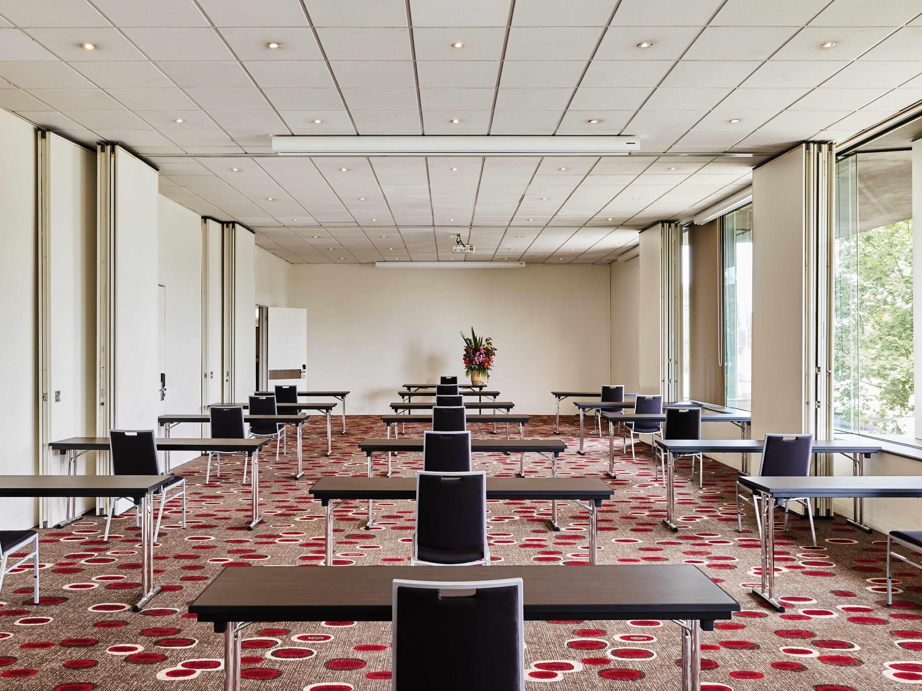 A conference room with tables and chairs, a carpet with a red and white pattern, a projector screen, and large windows.