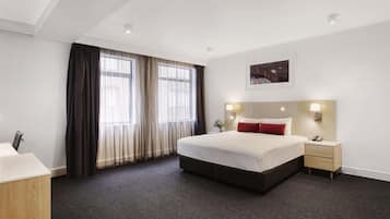A hotel room with a large bed, a desk, a chair, a nightstand, and a window with curtains.