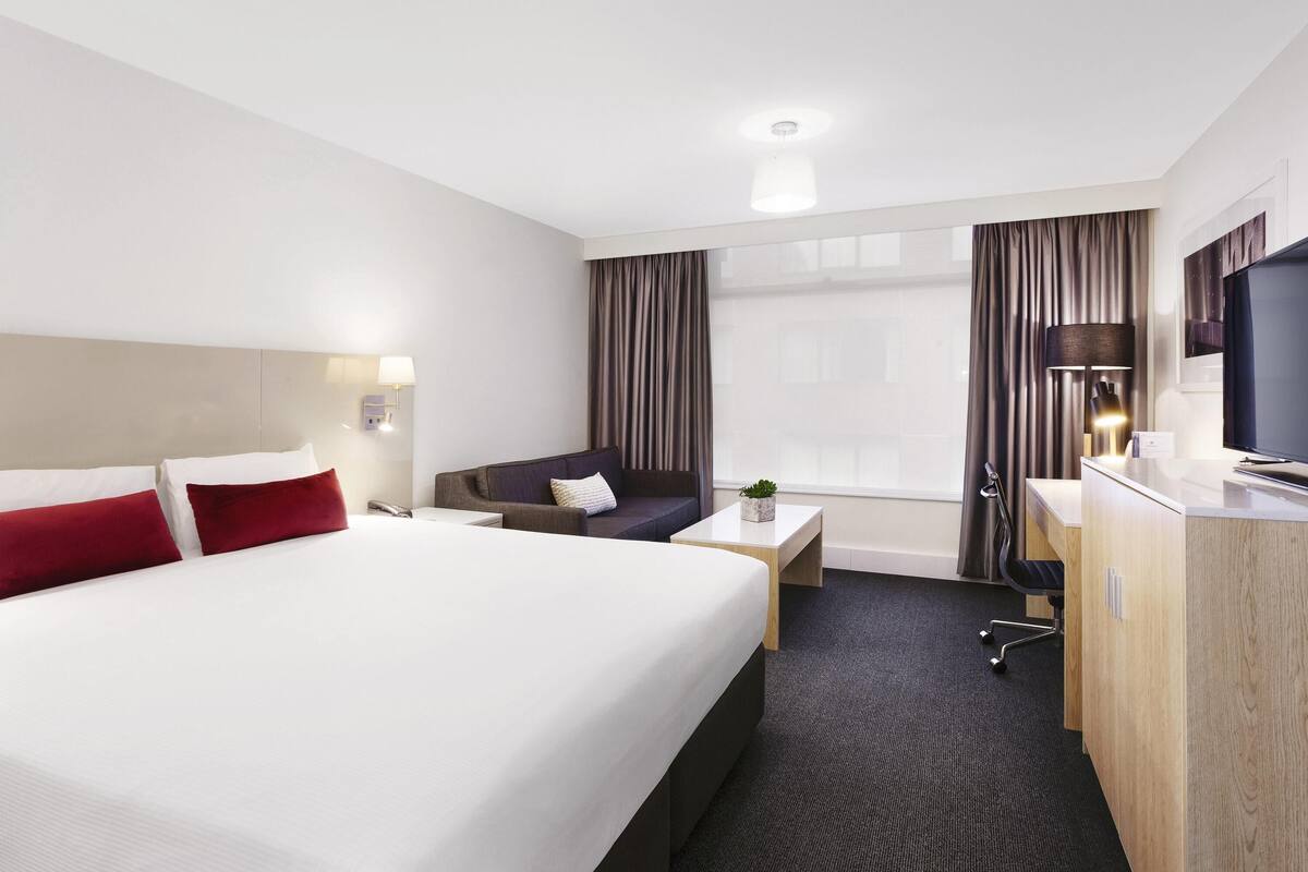 Une chambre d’hôtel avec un grand lit, un bureau, une chaise, une télévision et une fenêtre avec des rideaux.
