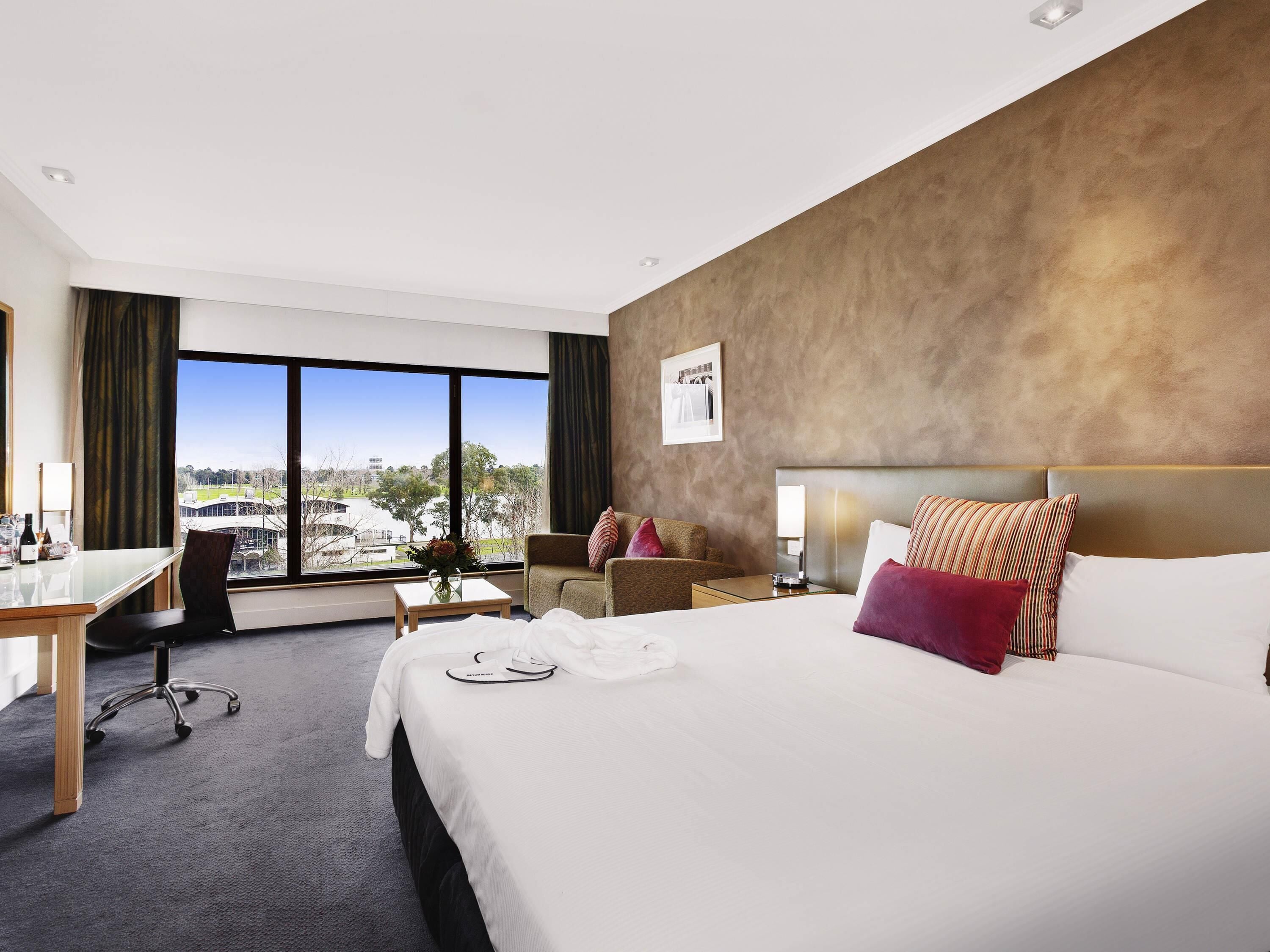 A hotel room with a large bed, a desk, a chair, and a view of a sports field.