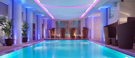 A modern indoor swimming pool with a central island, surrounded by lounge chairs and potted plants.