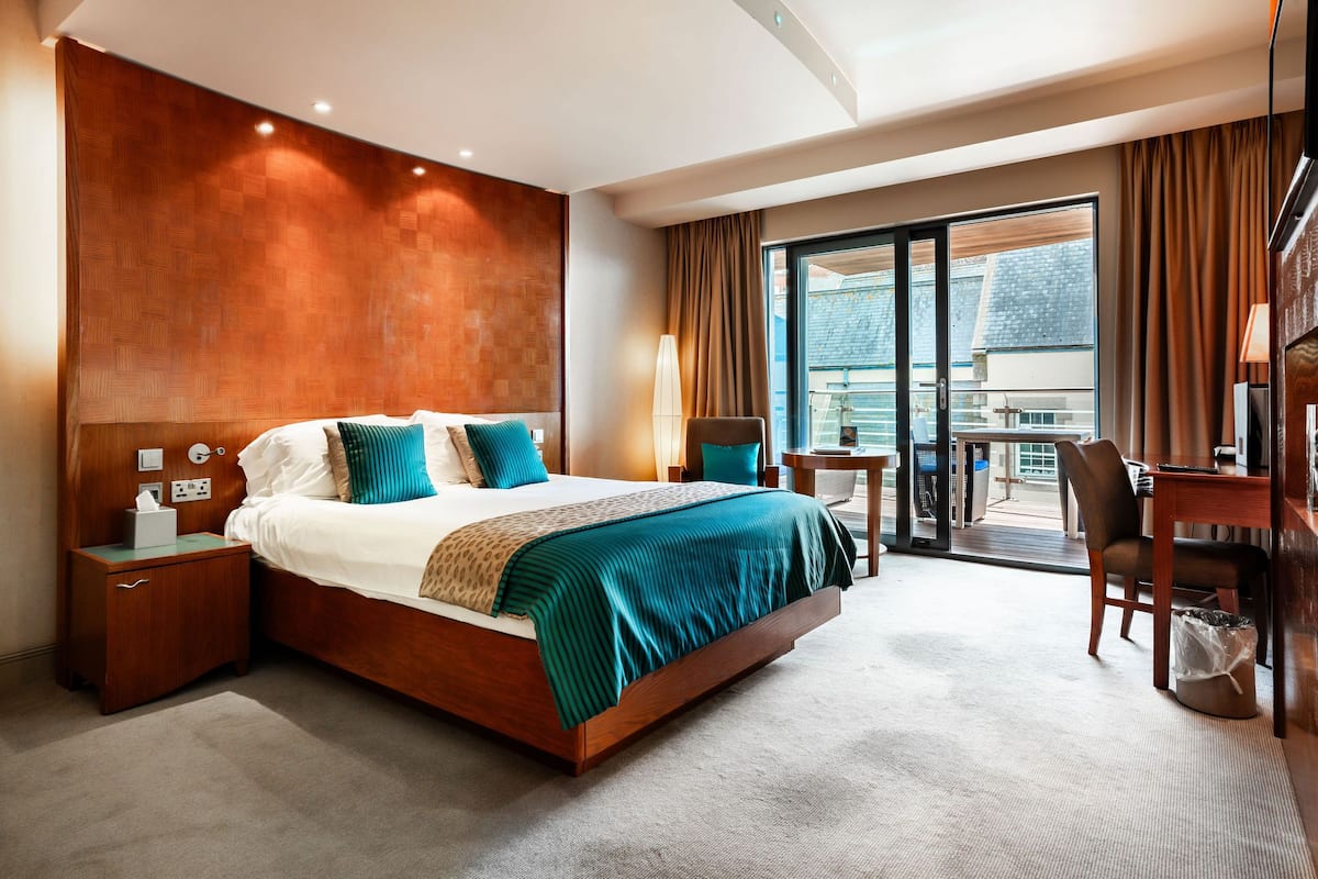 A hotel room with a large bed, a desk, a chair, a small table, and a balcony with a view.