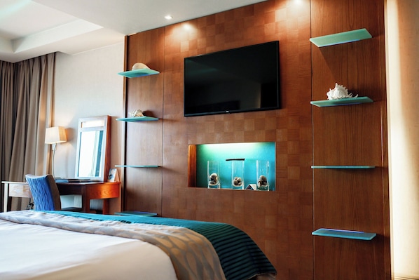Habitación de hotel con una cama grande, un escritorio con silla, un televisor de pantalla plana en la pared y un estante con objetos decorativos.