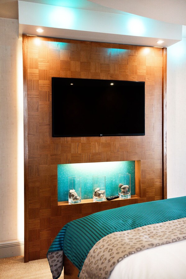 A hotel room with a wooden wall panel, a mounted TV, and a shelf with decorative items. - The Royal Yacht (St. Helier)