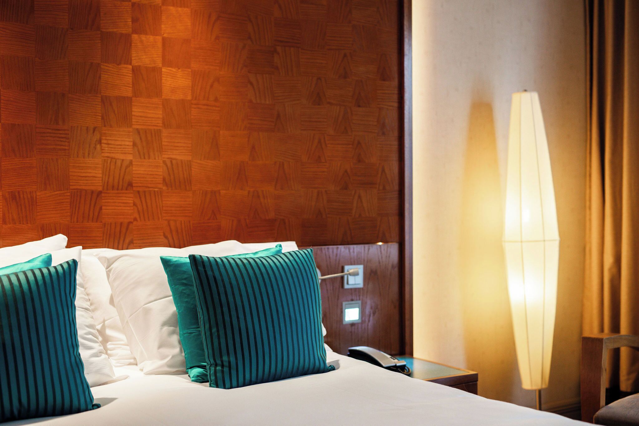 Una habitación de hotel con cabecera de madera, cama con sábanas blancas y almohadas color turquesa, una lámpara de noche y una mesita con un teléfono.
