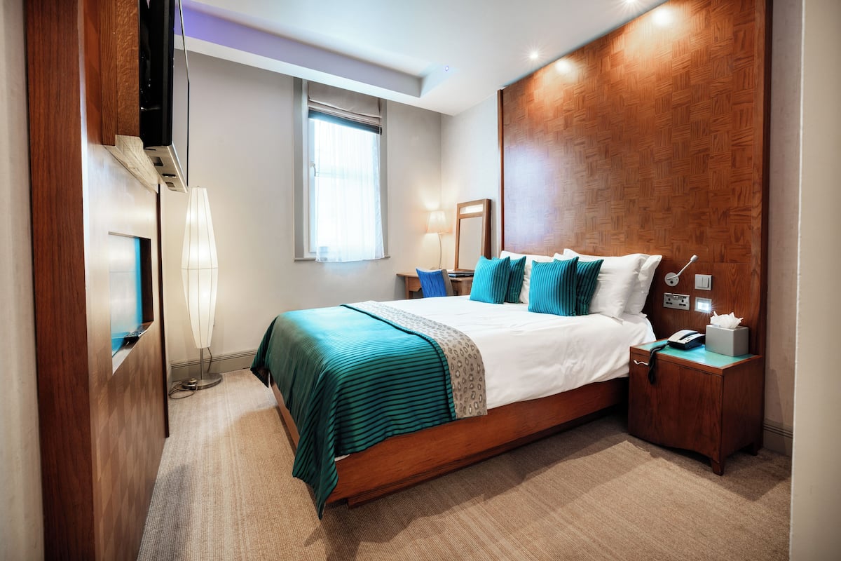 A modern hotel room with a large bed, wooden headboard, bedside table, lamp, and a flat-screen TV.