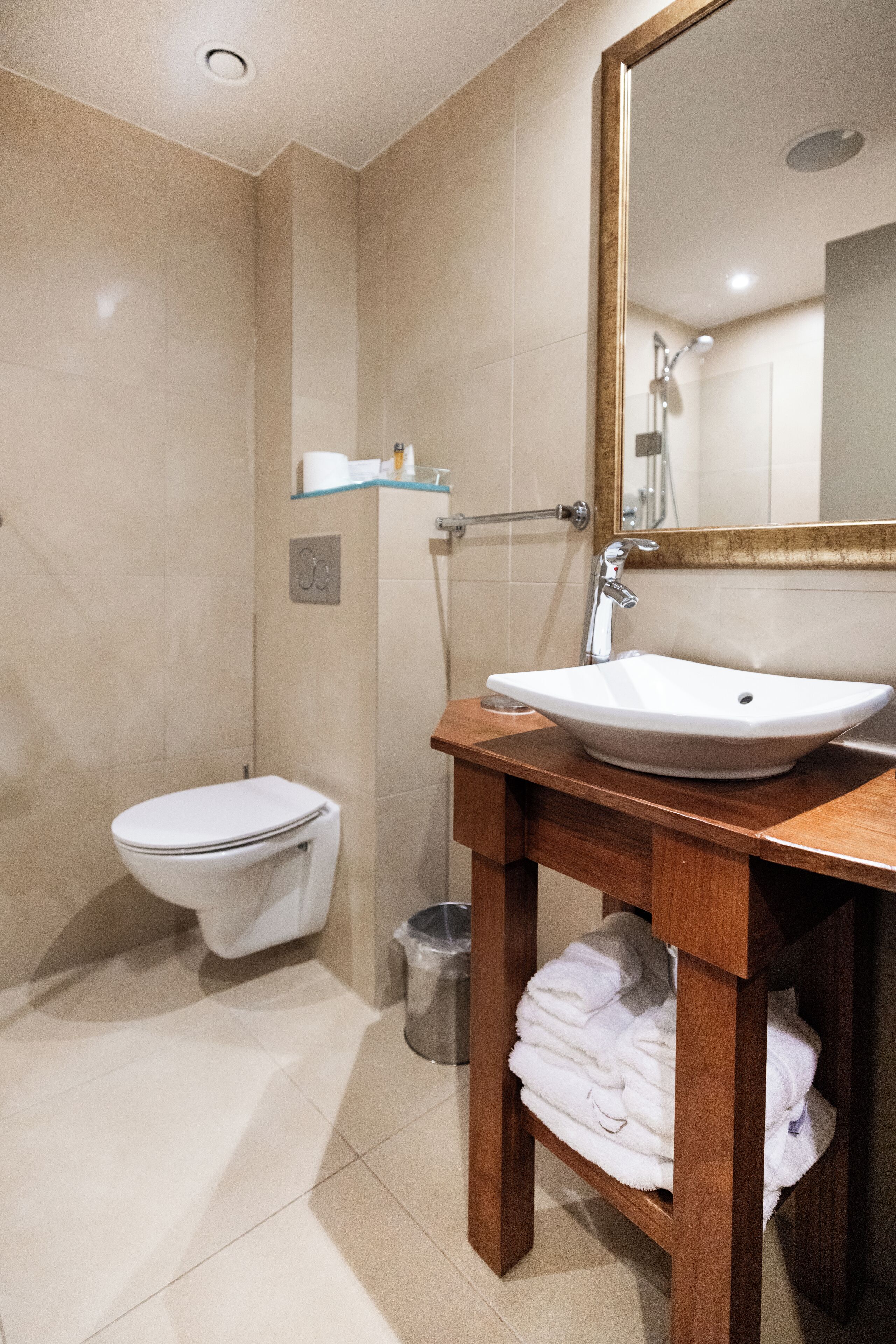 Un baño con lavamanos blanco, un espejo y un inodoro. Hay un perchero y un cubo de basura.