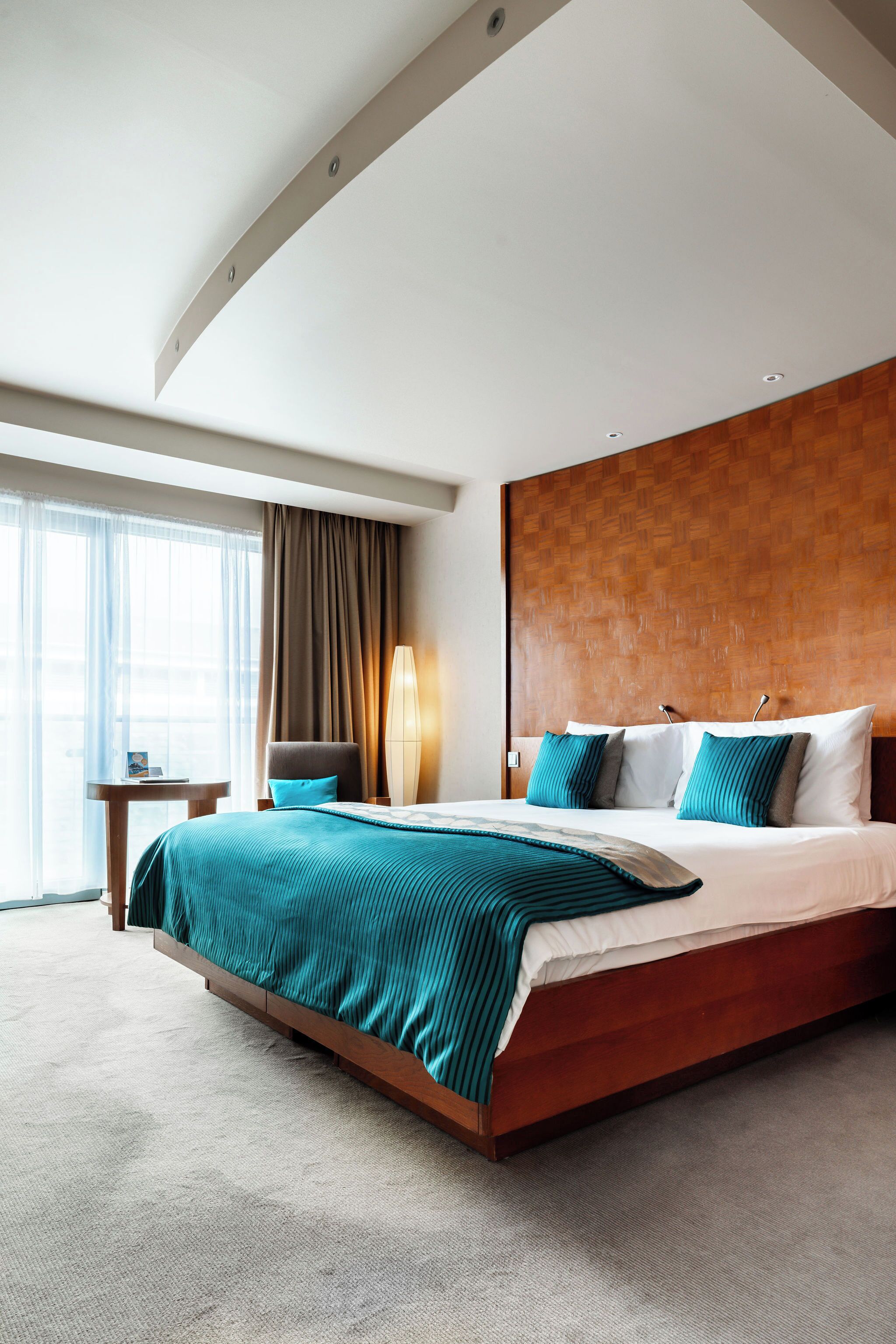 A hotel room with a large bed, a wooden headboard, a bedside table, a lamp, and a view of the outdoors through a window with curtains.