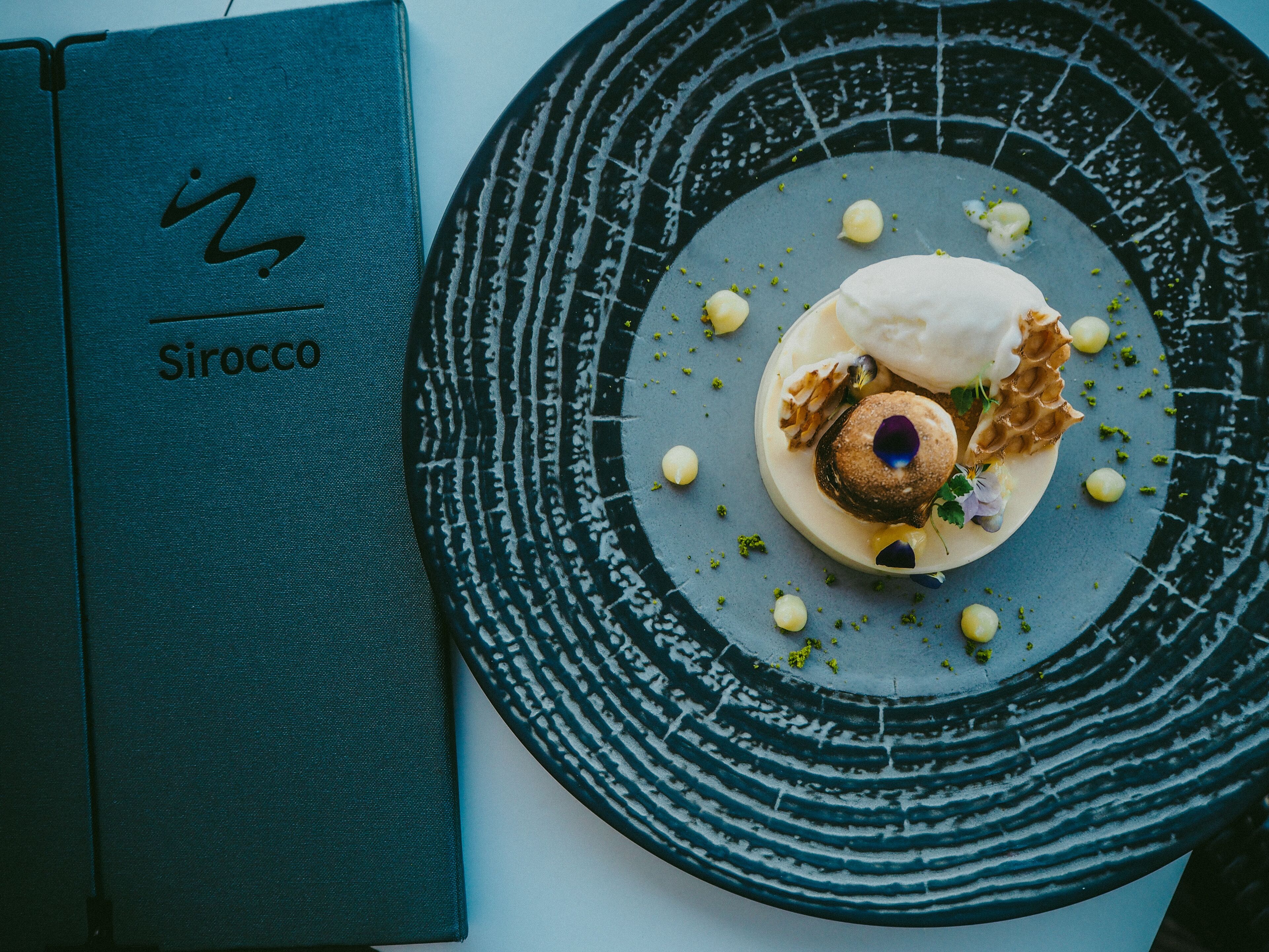 Un menú titulado 'Sirocco' sobre un fondo azul aguamarina, un plato con un postre que incluye un gofre, una bola de helado y guarniciones, y un plato oscuro y con textura.