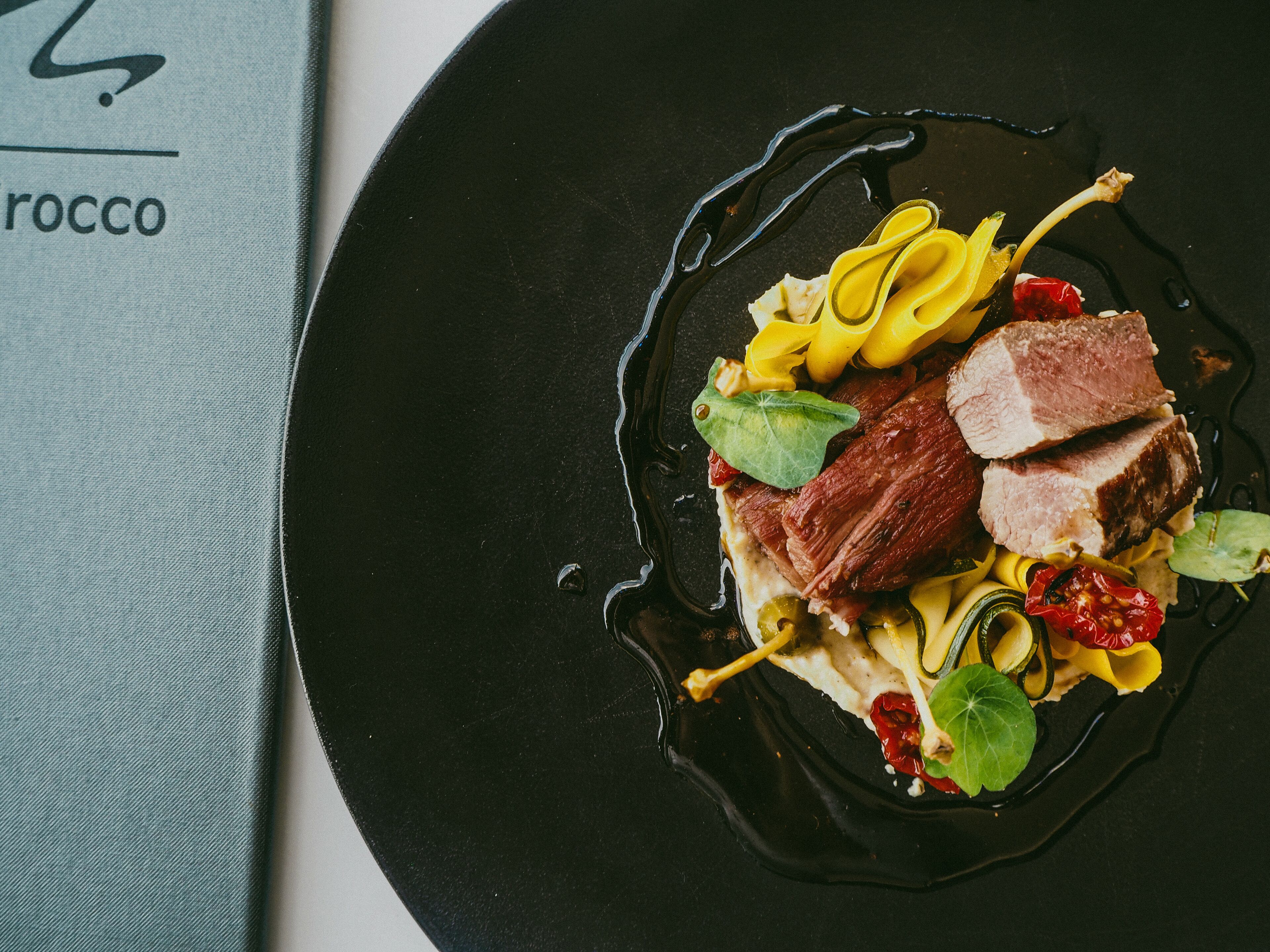 A black plate with a dish of food, including meat and vegetables, on a table.