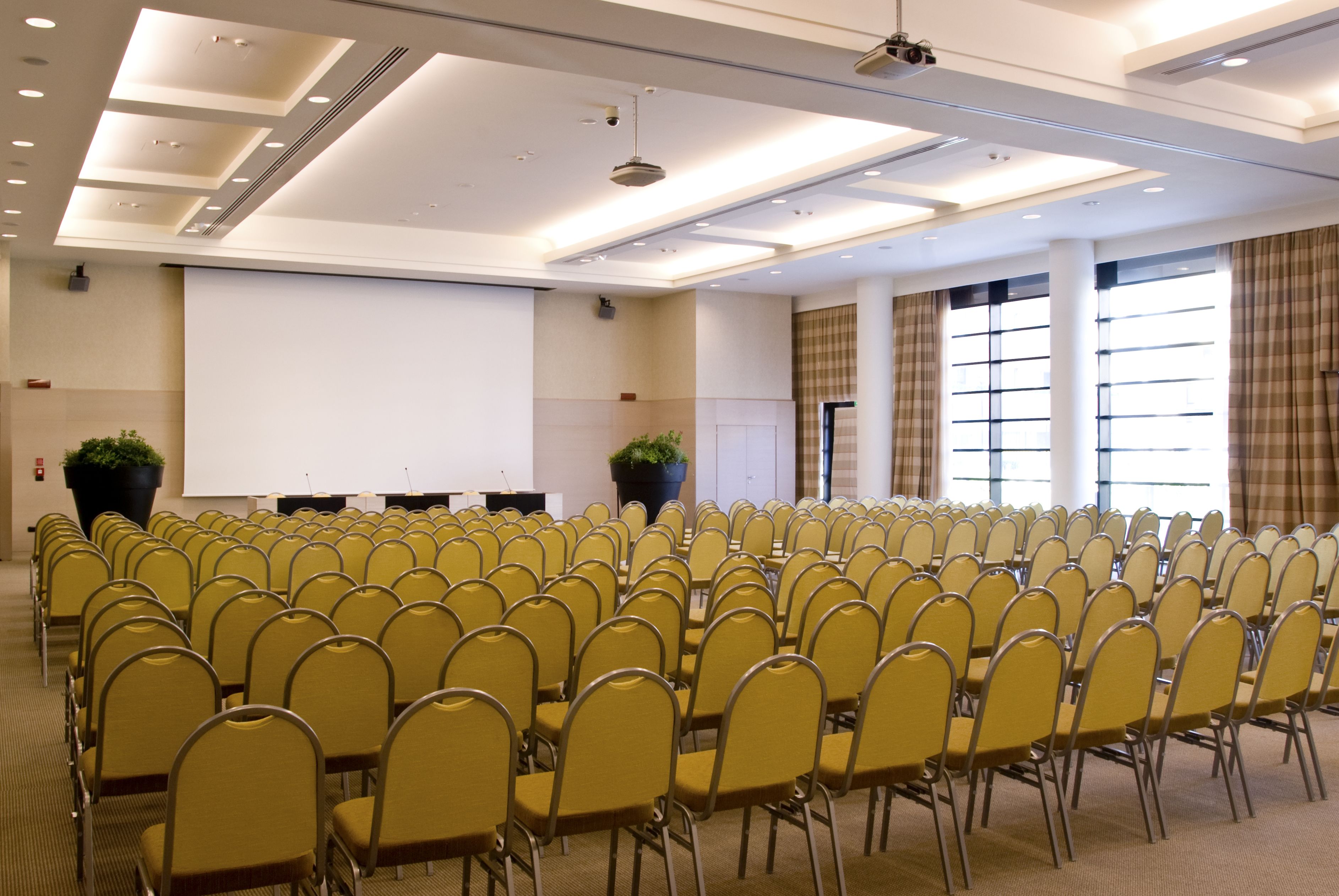 A conference room with rows of yellow chairs, a projector screen, and large windows with curtains.