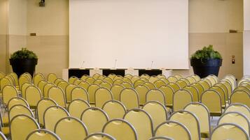 Una sala conferenze con file di sedie gialle, un grande schermo di proiezione e due piante in vaso.