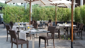 Un área de comedor al aire libre con mesas y sillas, sombrillas y follaje de fondo.