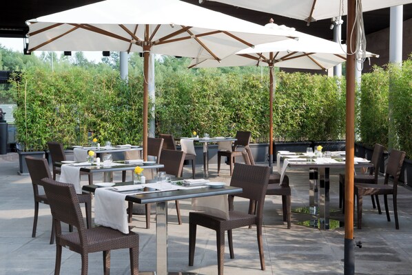 An outdoor dining area with tables and chairs, umbrellas, and greenery in the background. - Starhotels Grand Milan (Saronno)
