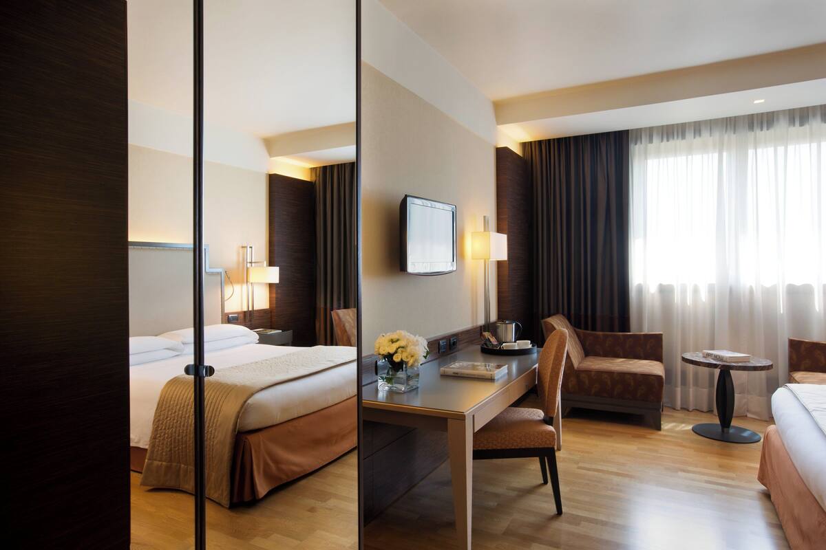 A hotel room with a bed, a desk, a chair, a small table, and a TV.
