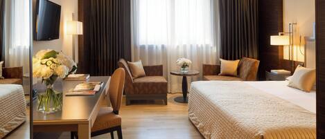 Habitación de hotel con cama, dos sillones, un escritorio con un florero y televisión. Ventana con cortinas.