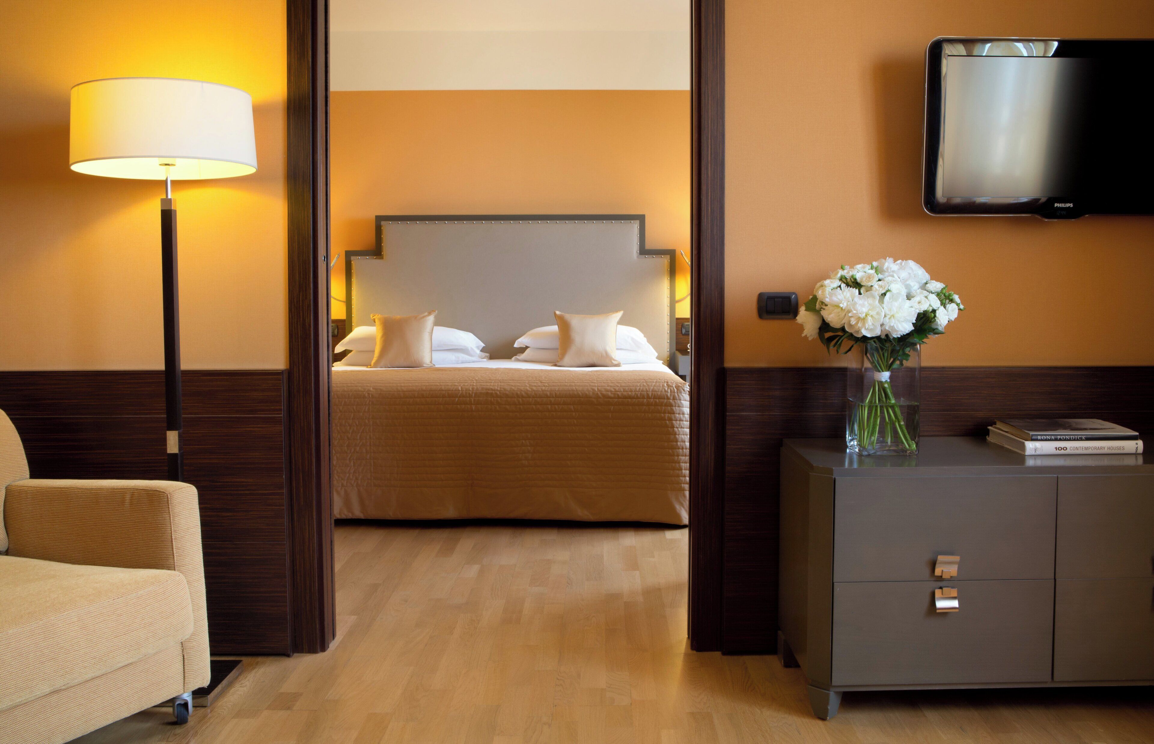 A hotel room with a bed, a bedside lamp, a TV, a vase with flowers, and a sofa.