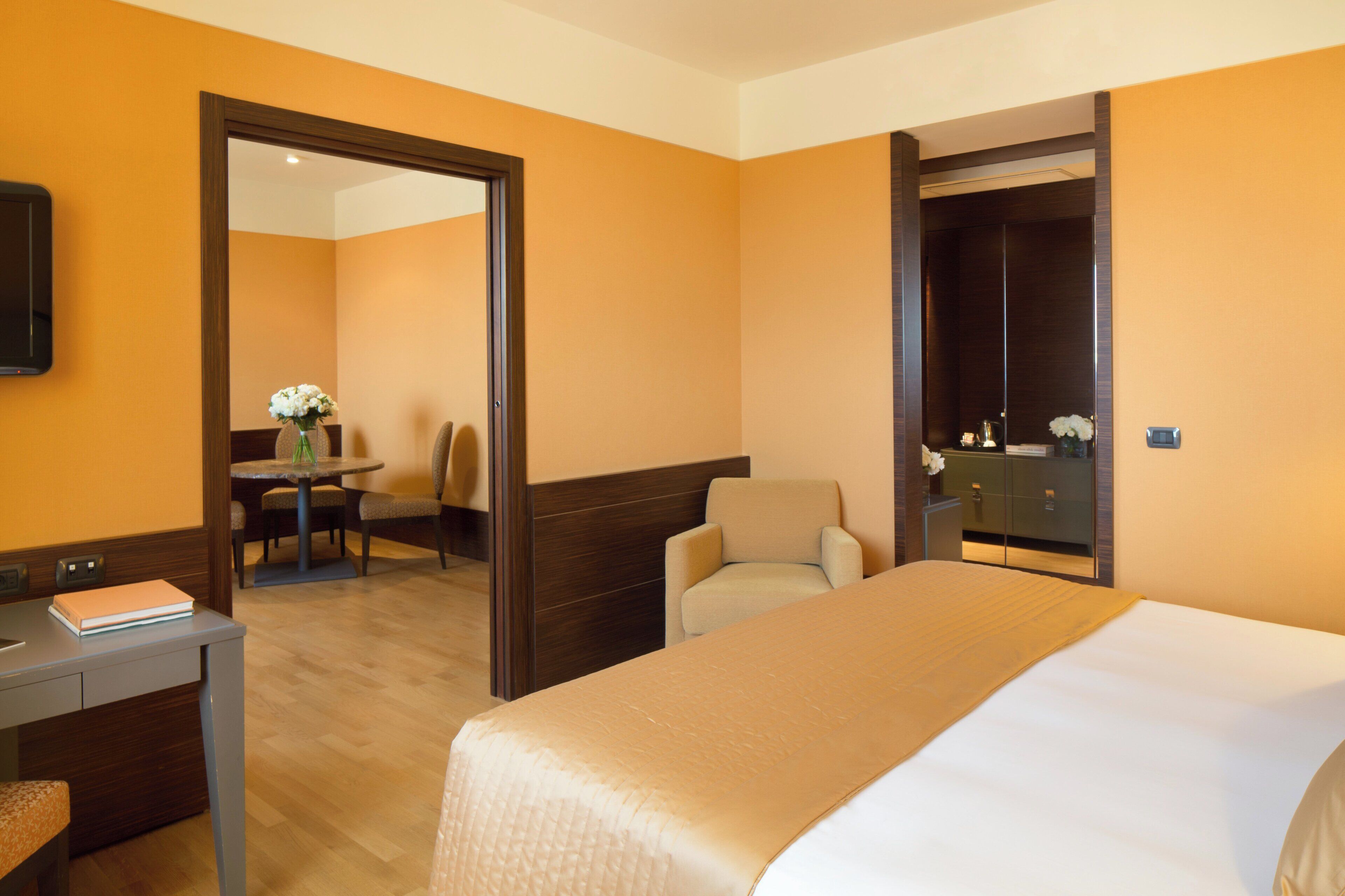 A hotel room with a bed, a desk, a chair, a table with flowers, and a mirror.