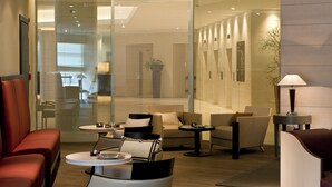 Un moderno vestíbulo de hotel con una zona de estar que incluye sofás y sillones, una mesita con lámpara, y un separador de vidrio con persianas.