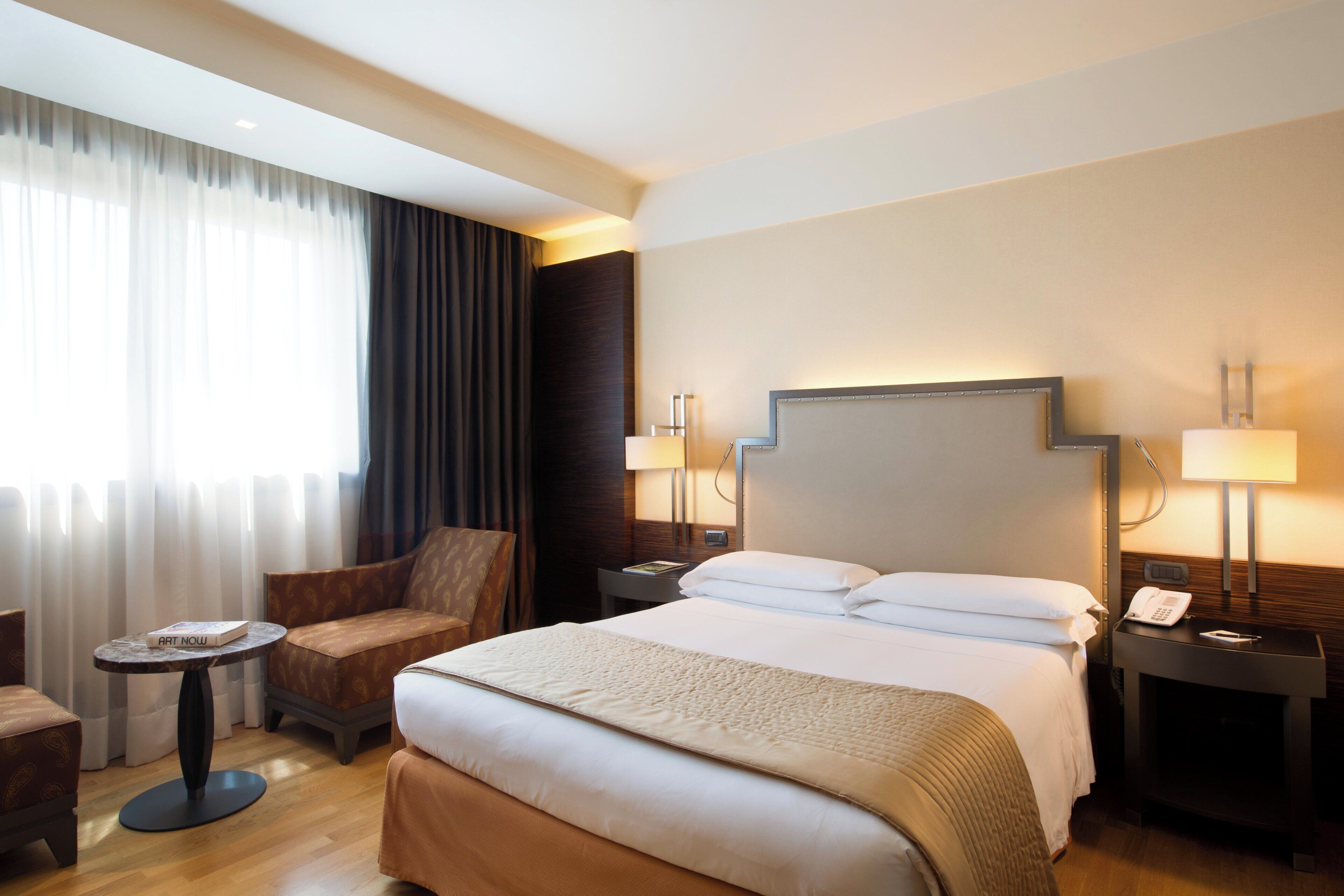 A hotel room with a large bed, two bedside tables with lamps, a small round table with a book, a sofa, and a window with curtains.